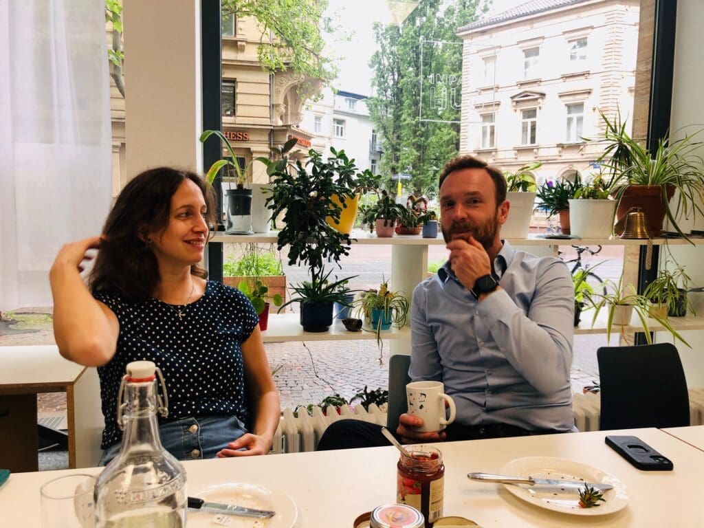 2 Mesnchen sitzen am Tisch, dahinter der Schaufensterblick auf die Fußgängerzone. Sie trinken Kaffee